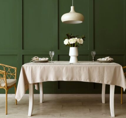 An elegant, eye-level wide shot of a dining area featuring a deep pepper tree green wainscoted wall. Centred in the room is a rectangular white wooden dining table draped with a light beige tablecloth, set with two minimalist place settings, crystal wine glasses, and a white vase holding white ranunculus flowers.