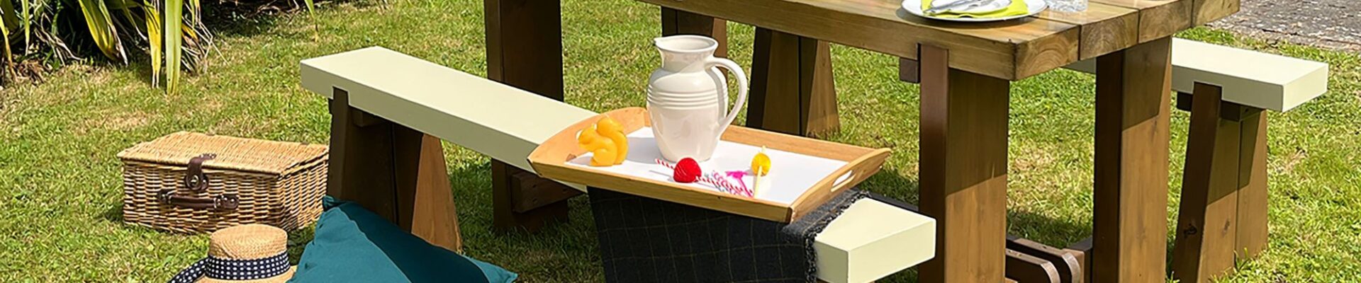 An outdoor wooden dining set sits on a lush green lawn on a sunny day. The set features a heavy, stained wooden table and two matching benches with contrasting cream-colored tops.