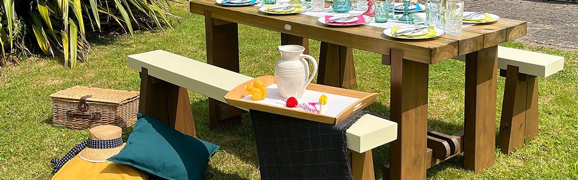 An outdoor wooden dining set sits on a lush green lawn on a sunny day. The set features a heavy, stained wooden table and two matching benches with contrasting cream-colored tops.