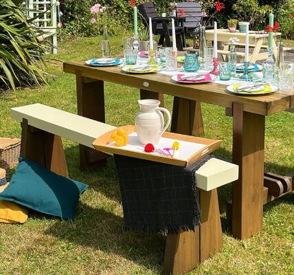 An outdoor wooden dining set sits on a lush green lawn on a sunny day. The set features a heavy, stained wooden table and two matching benches with contrasting cream-colored tops.
