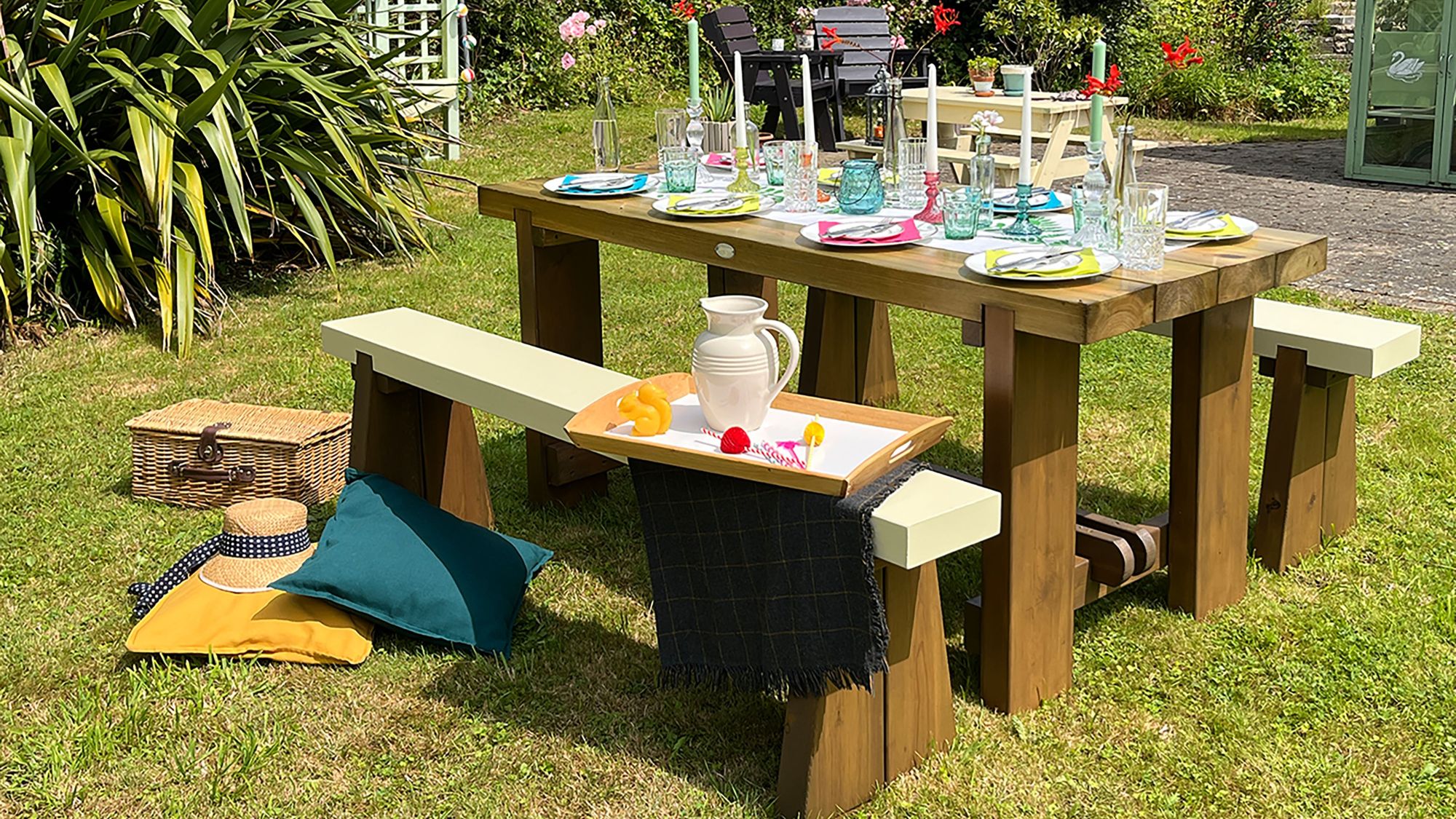 An outdoor wooden dining set sits on a lush green lawn on a sunny day. The set features a heavy, stained wooden table and two matching benches with contrasting cream-colored tops.