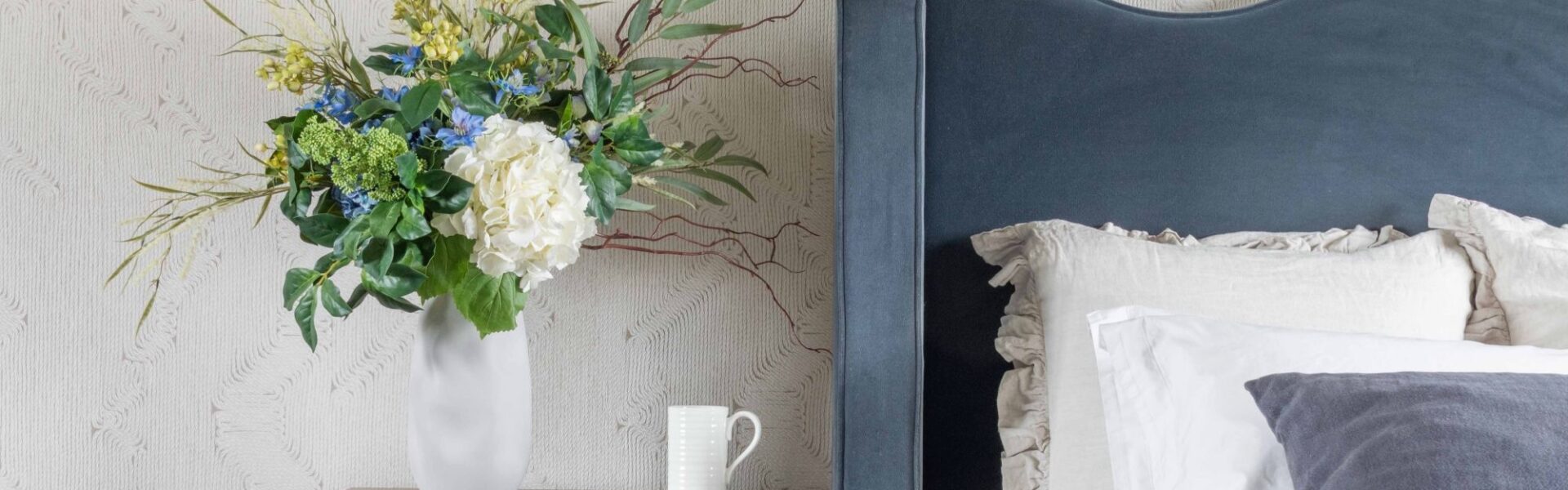 An elegant bedroom scene featuring a tall white organic-shaped ceramic vase filled with a lush artificial floral arrangement of white hydrangeas, blue delphiniums, and yellow wildflowers.