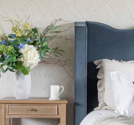 An elegant bedroom scene featuring a tall white organic-shaped ceramic vase filled with a lush artificial floral arrangement of white hydrangeas, blue delphiniums, and yellow wildflowers.