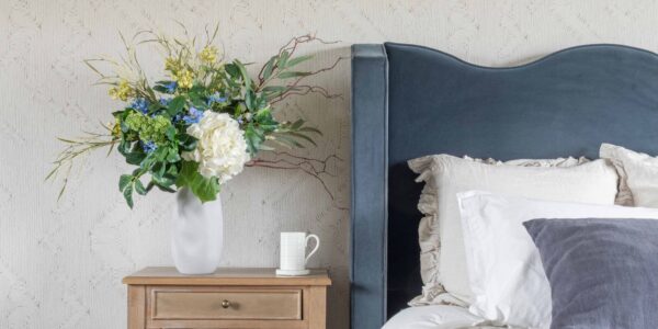 An elegant bedroom scene featuring a tall white organic-shaped ceramic vase filled with a lush artificial floral arrangement of white hydrangeas, blue delphiniums, and yellow wildflowers.