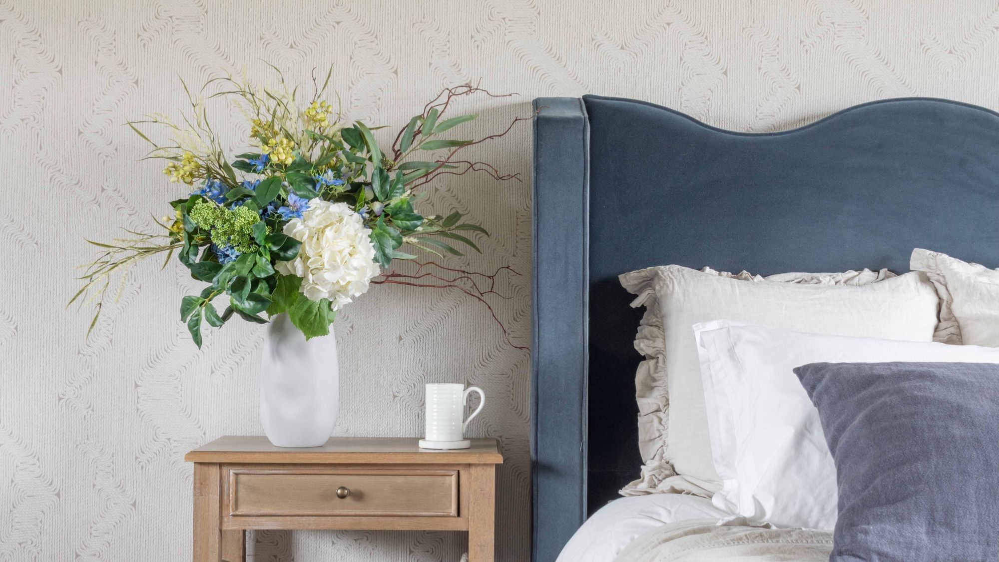 An elegant bedroom scene featuring a tall white organic-shaped ceramic vase filled with a lush artificial floral arrangement of white hydrangeas, blue delphiniums, and yellow wildflowers.
