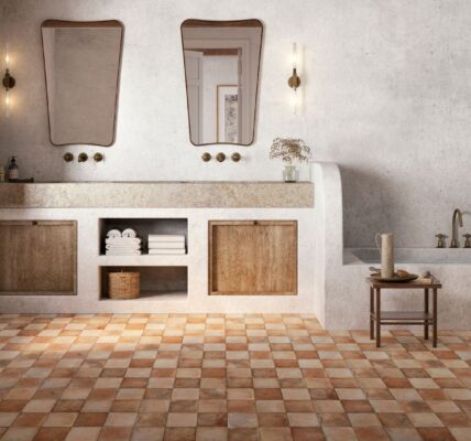 An interior photograph of a spacious, rustic-style bathroom featuring earthy tones and Mediterranean influences. The foreground is dominated by a chequerboard pattern of terracotta floor tiles in varying shades of burnt orange, tan, and cream.