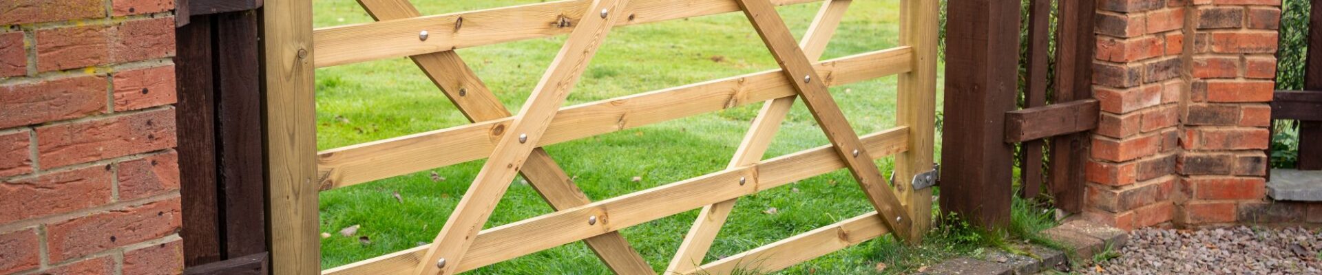 A 7ft light-coloured wooden five-bar gate installed between brick pillars, opening up to a green lawn in a countryside garden setting.