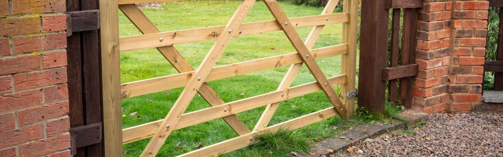 A 7ft light-coloured wooden five-bar gate installed between brick pillars, opening up to a green lawn in a countryside garden setting.
