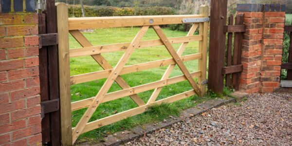 A 7ft light-coloured wooden five-bar gate installed between brick pillars, opening up to a green lawn in a countryside garden setting.