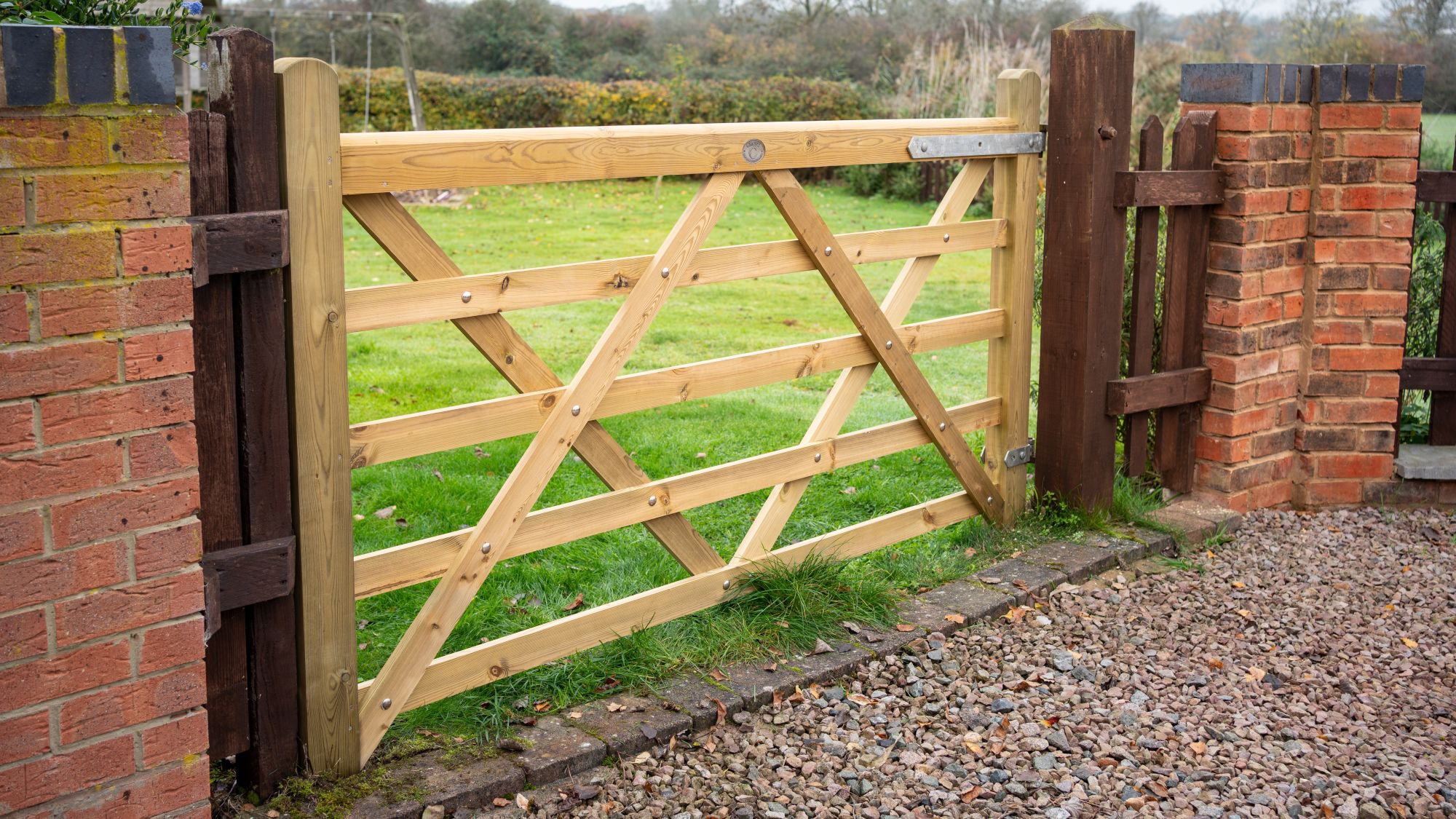 A 7ft light-coloured wooden five-bar gate installed between brick pillars, opening up to a green lawn in a countryside garden setting.