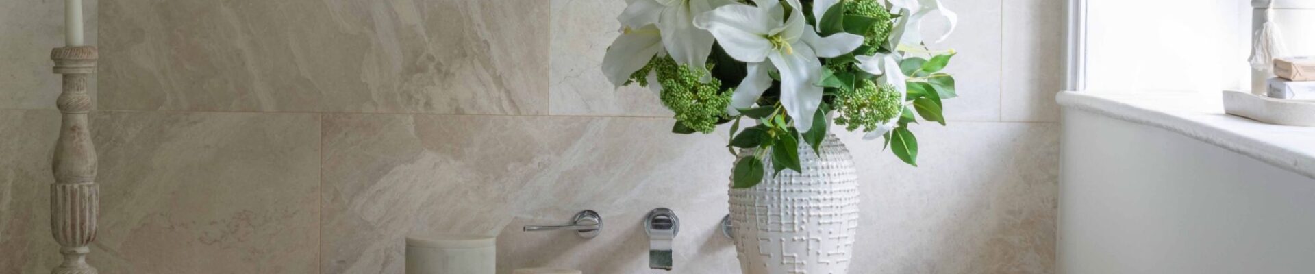 A serene, spa-like bathroom setting featuring a white freestanding bathtub positioned against a beige, natural stone tiled wall. A wooden bath caddy sits across the tub, holding three small ceramic canisters and a tall, textured white ceramic vase filled with a lush bouquet of white lilies and green foliage.