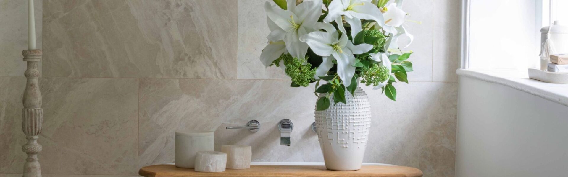 A serene, spa-like bathroom setting featuring a white freestanding bathtub positioned against a beige, natural stone tiled wall. A wooden bath caddy sits across the tub, holding three small ceramic canisters and a tall, textured white ceramic vase filled with a lush bouquet of white lilies and green foliage.