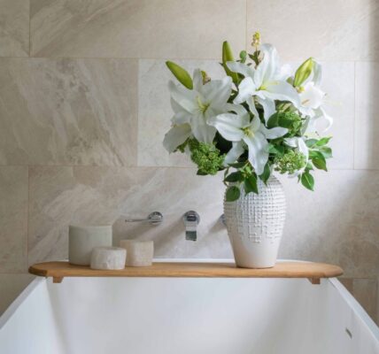 A serene, spa-like bathroom setting featuring a white freestanding bathtub positioned against a beige, natural stone tiled wall. A wooden bath caddy sits across the tub, holding three small ceramic canisters and a tall, textured white ceramic vase filled with a lush bouquet of white lilies and green foliage.