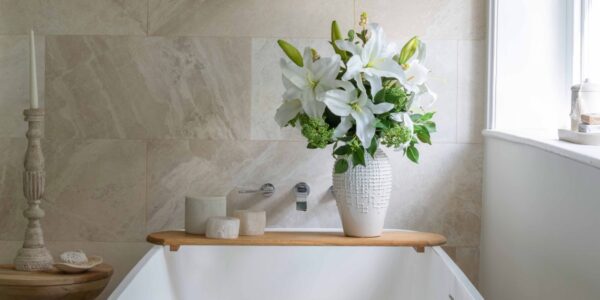 A serene, spa-like bathroom setting featuring a white freestanding bathtub positioned against a beige, natural stone tiled wall. A wooden bath caddy sits across the tub, holding three small ceramic canisters and a tall, textured white ceramic vase filled with a lush bouquet of white lilies and green foliage.