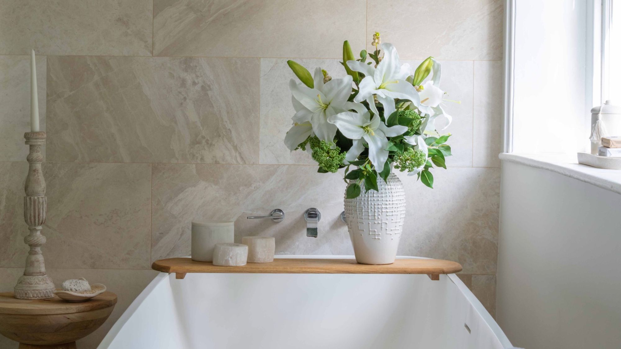 A serene, spa-like bathroom setting featuring a white freestanding bathtub positioned against a beige, natural stone tiled wall. A wooden bath caddy sits across the tub, holding three small ceramic canisters and a tall, textured white ceramic vase filled with a lush bouquet of white lilies and green foliage.