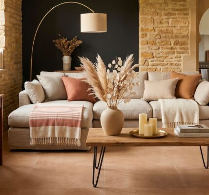 A warm, rustic living room featuring a light beige sectional sofa, a wooden coffee table with hairpin legs, exposed stone walls, and terracotta floor tiles.
