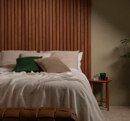 A cosy, modern bedroom featuring a bed with a large, warm-toned Sapele wood slat wall panel serving as a floor-to-ceiling headboard.