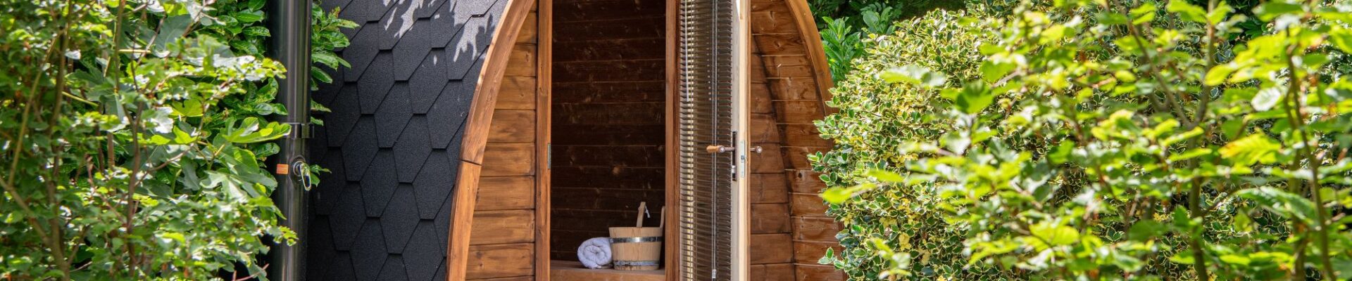 An outdoor, barrel-arched wooden sauna nestled in a lush green garden setting. The sauna features a dark, hexagonal-shingled roof and an open glass door equipped with horizontal blinds, revealing a wooden bench, a rolled white towel, and a traditional sauna bucket inside.