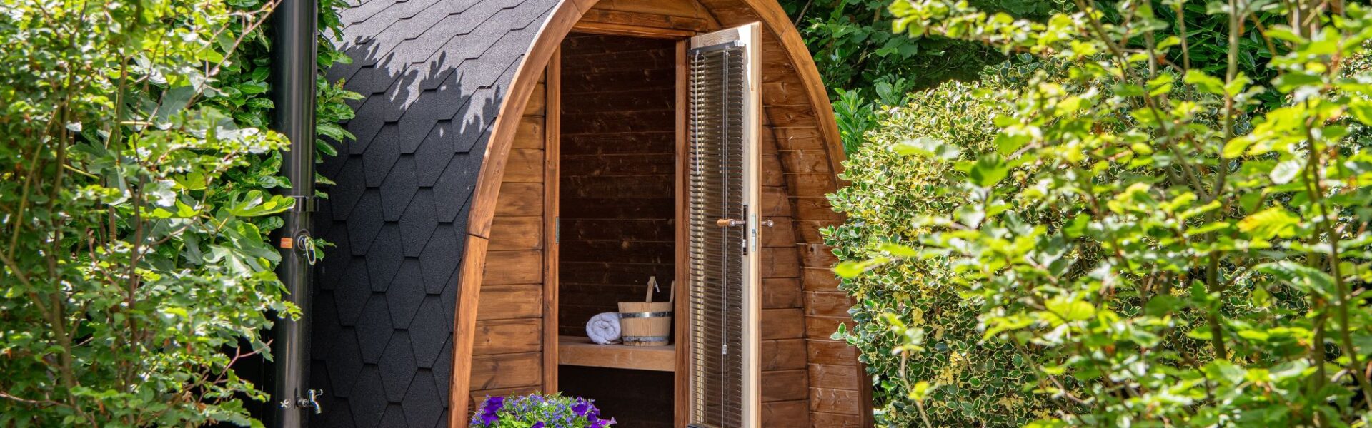 An outdoor, barrel-arched wooden sauna nestled in a lush green garden setting. The sauna features a dark, hexagonal-shingled roof and an open glass door equipped with horizontal blinds, revealing a wooden bench, a rolled white towel, and a traditional sauna bucket inside.