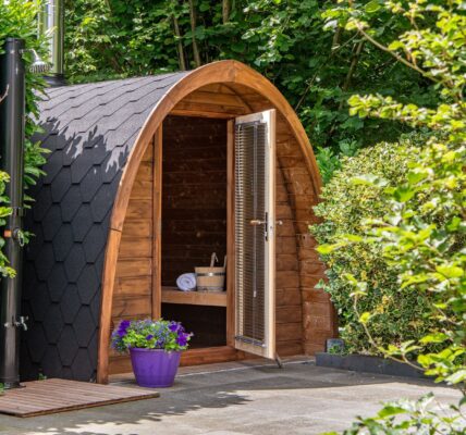 An outdoor, barrel-arched wooden sauna nestled in a lush green garden setting. The sauna features a dark, hexagonal-shingled roof and an open glass door equipped with horizontal blinds, revealing a wooden bench, a rolled white towel, and a traditional sauna bucket inside.