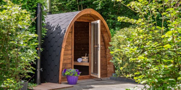 An outdoor, barrel-arched wooden sauna nestled in a lush green garden setting. The sauna features a dark, hexagonal-shingled roof and an open glass door equipped with horizontal blinds, revealing a wooden bench, a rolled white towel, and a traditional sauna bucket inside.