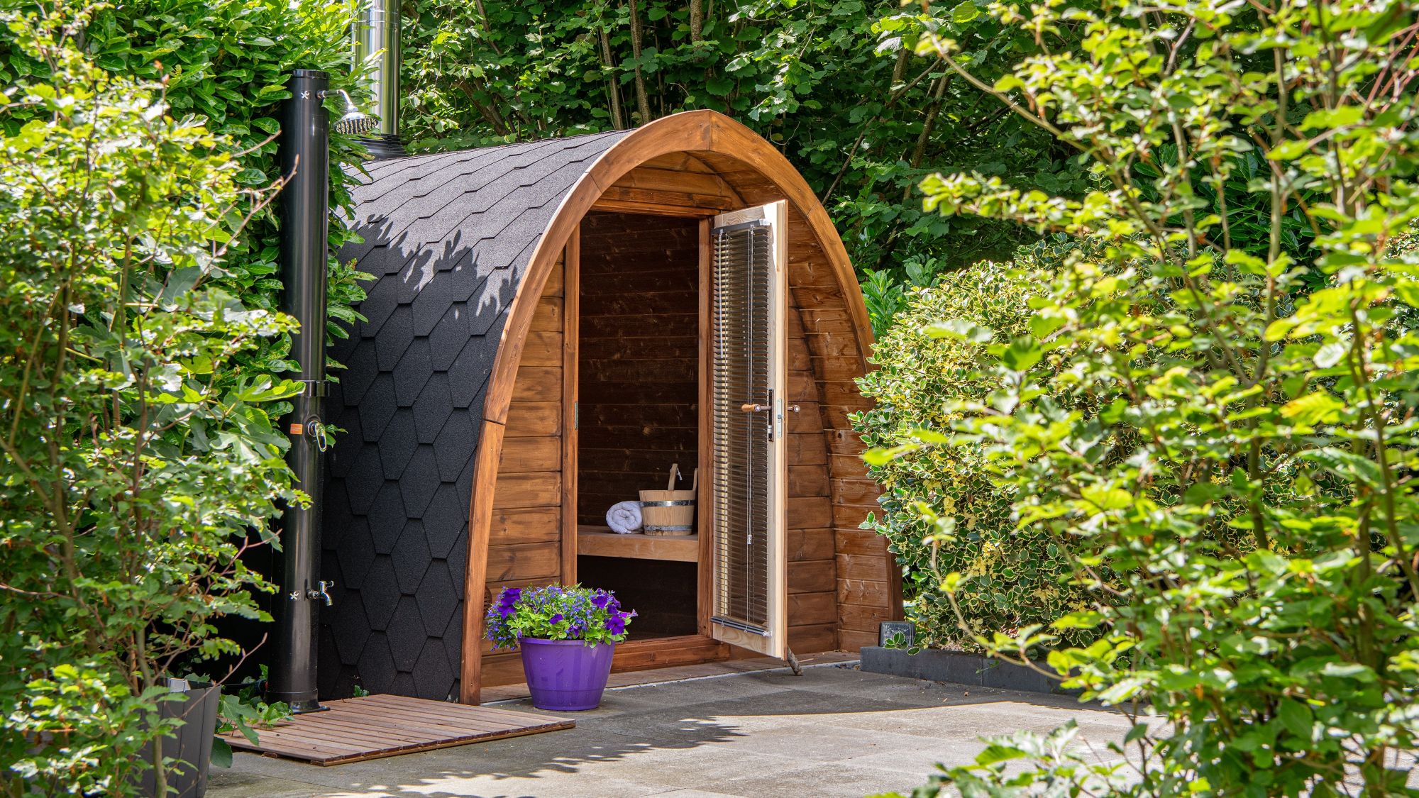 An outdoor, barrel-arched wooden sauna nestled in a lush green garden setting. The sauna features a dark, hexagonal-shingled roof and an open glass door equipped with horizontal blinds, revealing a wooden bench, a rolled white towel, and a traditional sauna bucket inside.