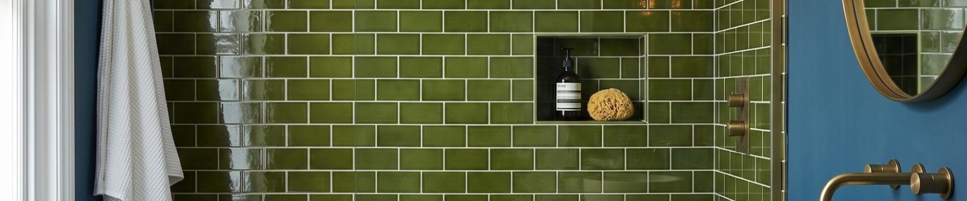 A modern bathroom featuring a shower clad in glossy olive green subway tiles with a built-in niche holding a soap bottle and sponge. To the right, a round stone vessel sink sits on a blue vanity next to brass hardware, a lit candle, and a round mirror reflecting a double-bulb wall sconce.