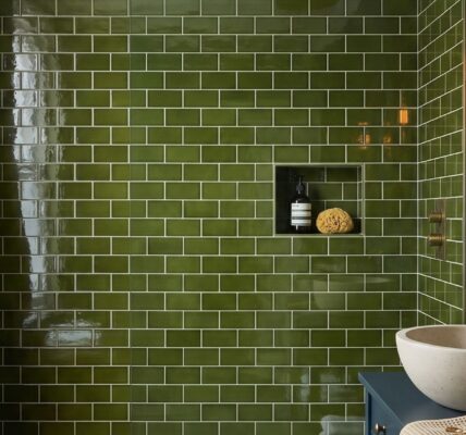 A modern bathroom featuring a shower clad in glossy olive green subway tiles with a built-in niche holding a soap bottle and sponge. To the right, a round stone vessel sink sits on a blue vanity next to brass hardware, a lit candle, and a round mirror reflecting a double-bulb wall sconce.