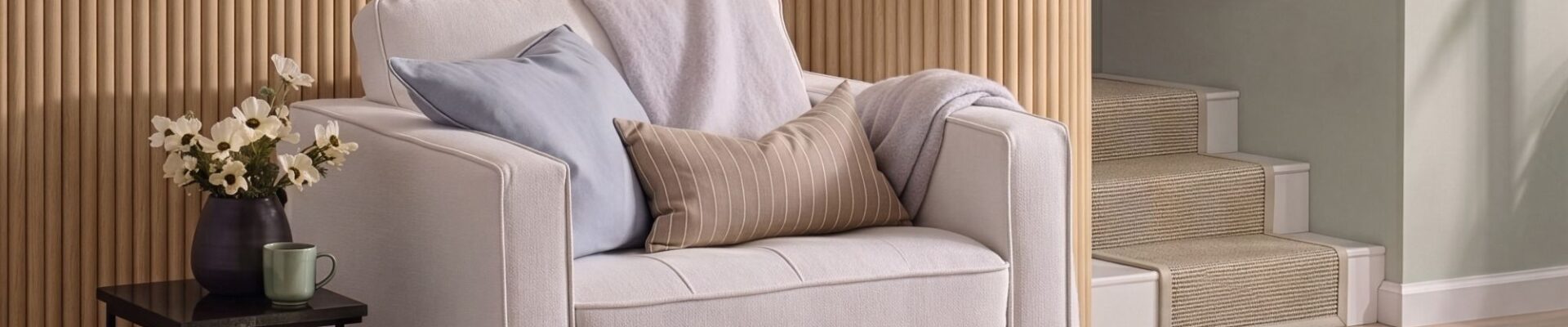 A cosy, modern white armchair sits in front of a light oak reeded wood-panelled feature wall. The armchair is styled with a light blue cushion, a tan striped lumbar pillow, and a soft lavender throw blanket.