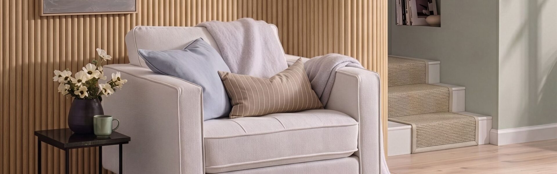 A cosy, modern white armchair sits in front of a light oak reeded wood-panelled feature wall. The armchair is styled with a light blue cushion, a tan striped lumbar pillow, and a soft lavender throw blanket.