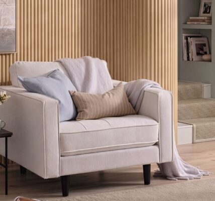 A cosy, modern white armchair sits in front of a light oak reeded wood-panelled feature wall. The armchair is styled with a light blue cushion, a tan striped lumbar pillow, and a soft lavender throw blanket.