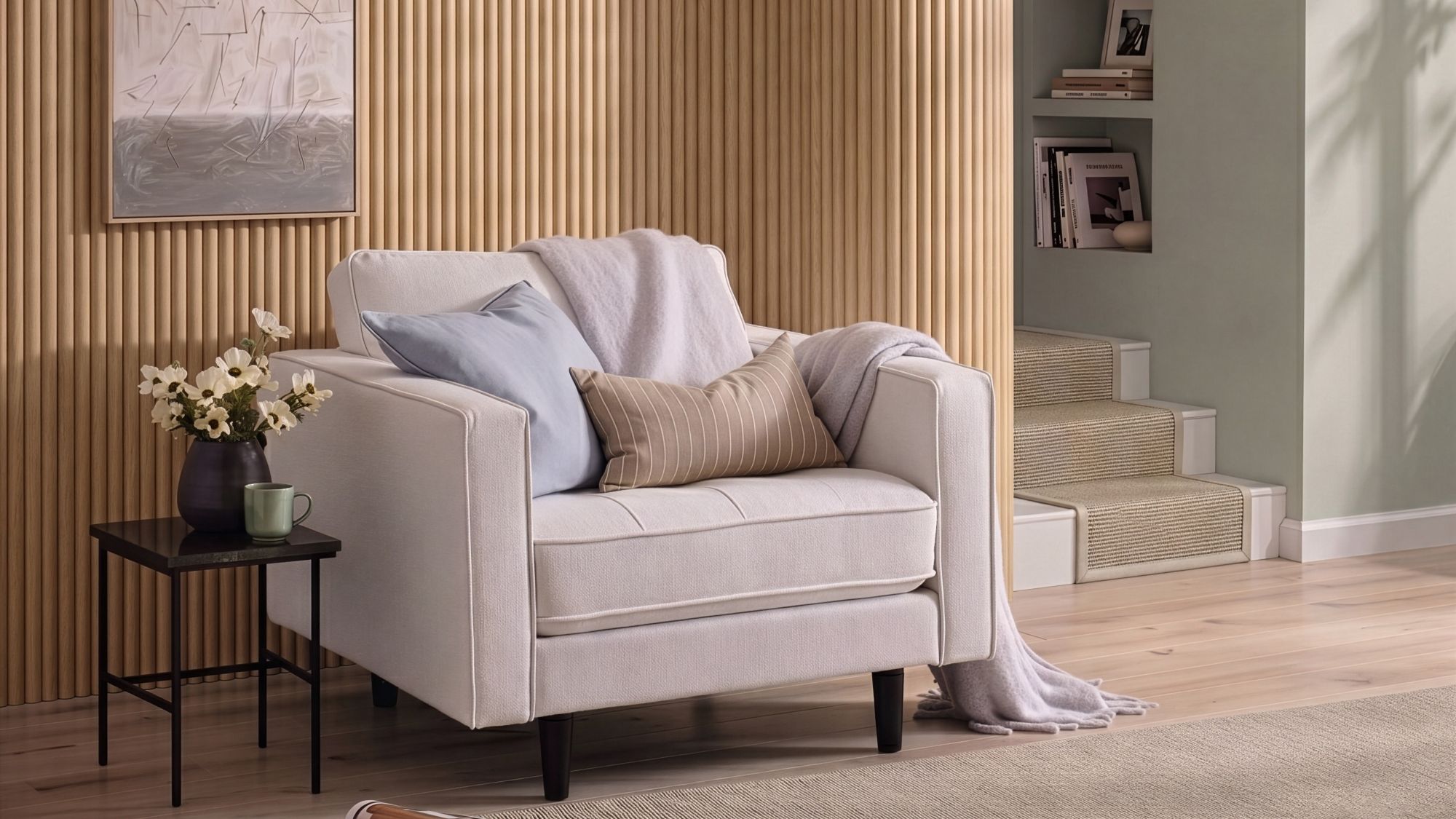 A cosy, modern white armchair sits in front of a light oak reeded wood-panelled feature wall. The armchair is styled with a light blue cushion, a tan striped lumbar pillow, and a soft lavender throw blanket.