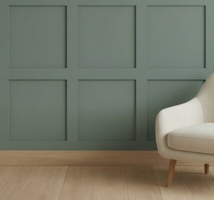 A sage green wall featuring a classic square Shaker-style wood panelling kit. A modern cream upholstered armchair with light wood legs sits on a light oak wood floor in front of the panelled wall.