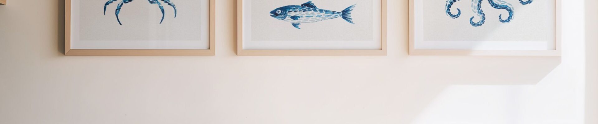 Three square, light-wood framed art pieces hang on a neutral beige wall above a kitchen countertop. Each frame features a watercolor-style illustration in shades of blue against a white background, depicting marine life: a crab on the left, three stacked fish in the center, and an octopus on the right.