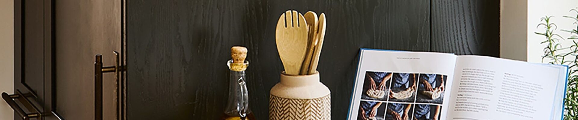 A kitchen counter styled with an open cookbook displaying bread-making steps, a bottle of olive oil, wooden utensils in a ceramic holder, and small condiment bowls against a dark wooden cabinet backdrop.
