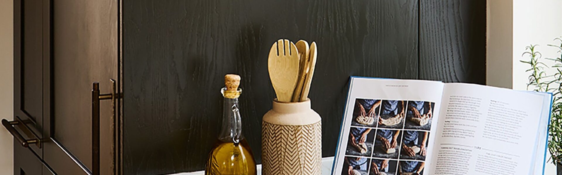 A kitchen counter styled with an open cookbook displaying bread-making steps, a bottle of olive oil, wooden utensils in a ceramic holder, and small condiment bowls against a dark wooden cabinet backdrop.