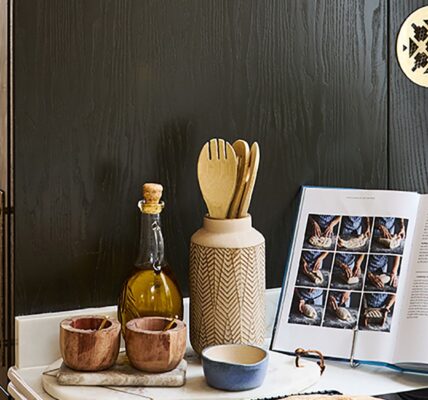 A kitchen counter styled with an open cookbook displaying bread-making steps, a bottle of olive oil, wooden utensils in a ceramic holder, and small condiment bowls against a dark wooden cabinet backdrop.