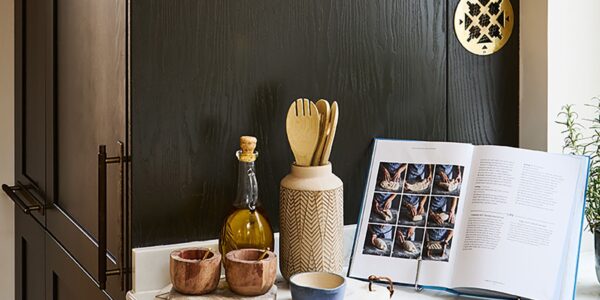 A kitchen counter styled with an open cookbook displaying bread-making steps, a bottle of olive oil, wooden utensils in a ceramic holder, and small condiment bowls against a dark wooden cabinet backdrop.