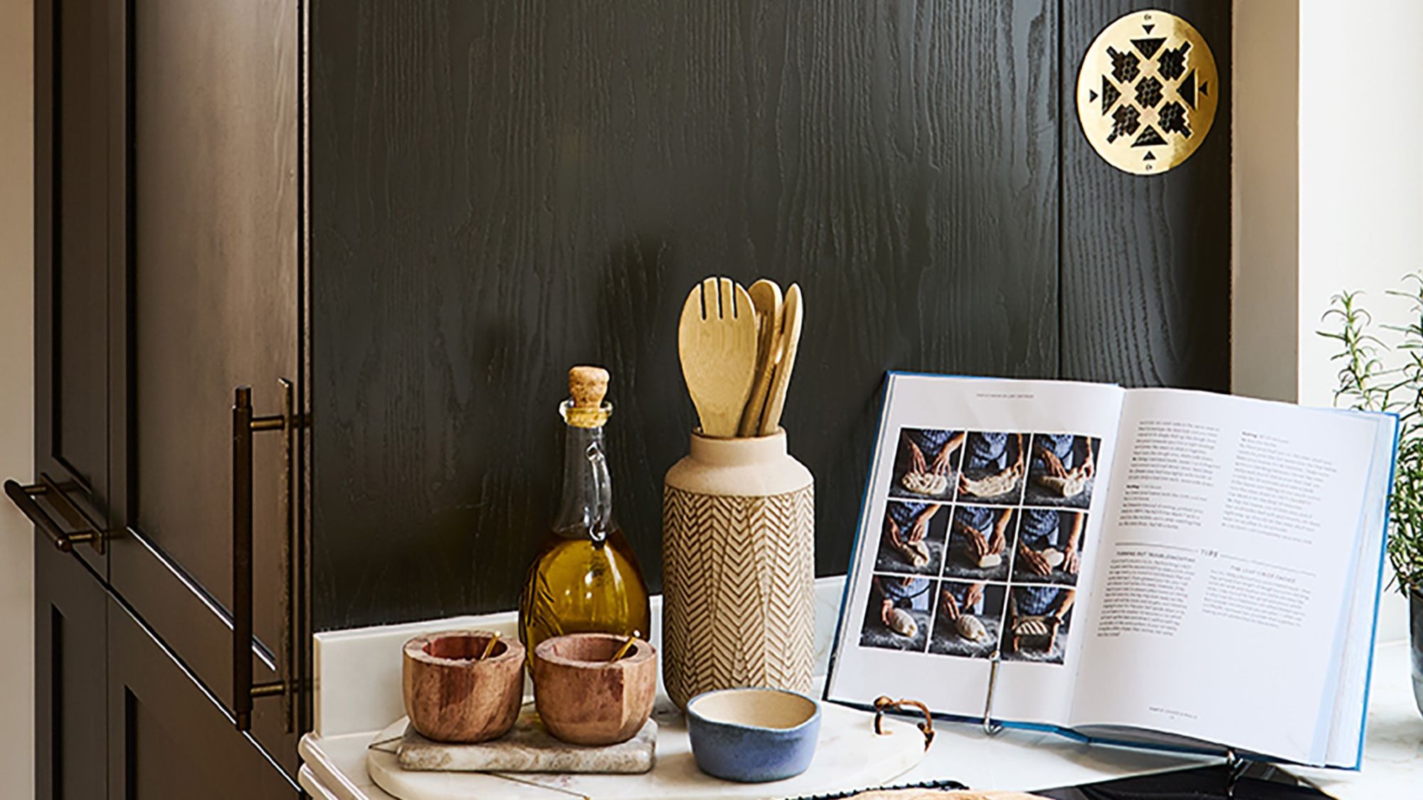 A kitchen counter styled with an open cookbook displaying bread-making steps, a bottle of olive oil, wooden utensils in a ceramic holder, and small condiment bowls against a dark wooden cabinet backdrop.