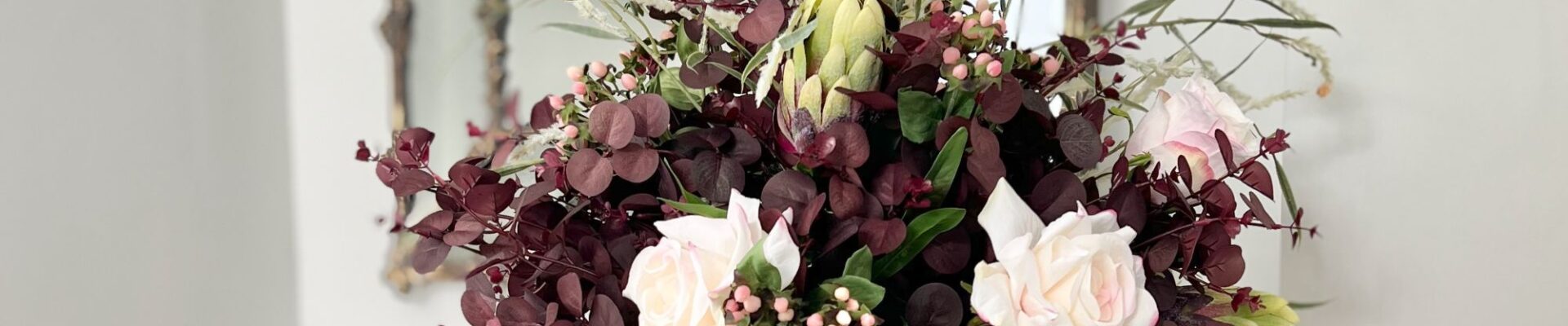 A lush autumn floral arrangement in a matte white ceramic vase, featuring deep plum-coloured eucalyptus foliage, pale pink roses, small pink berries, a green protea bud, and wispy ornamental grasses, set against a bright interior with a gold-framed mirror in the background.