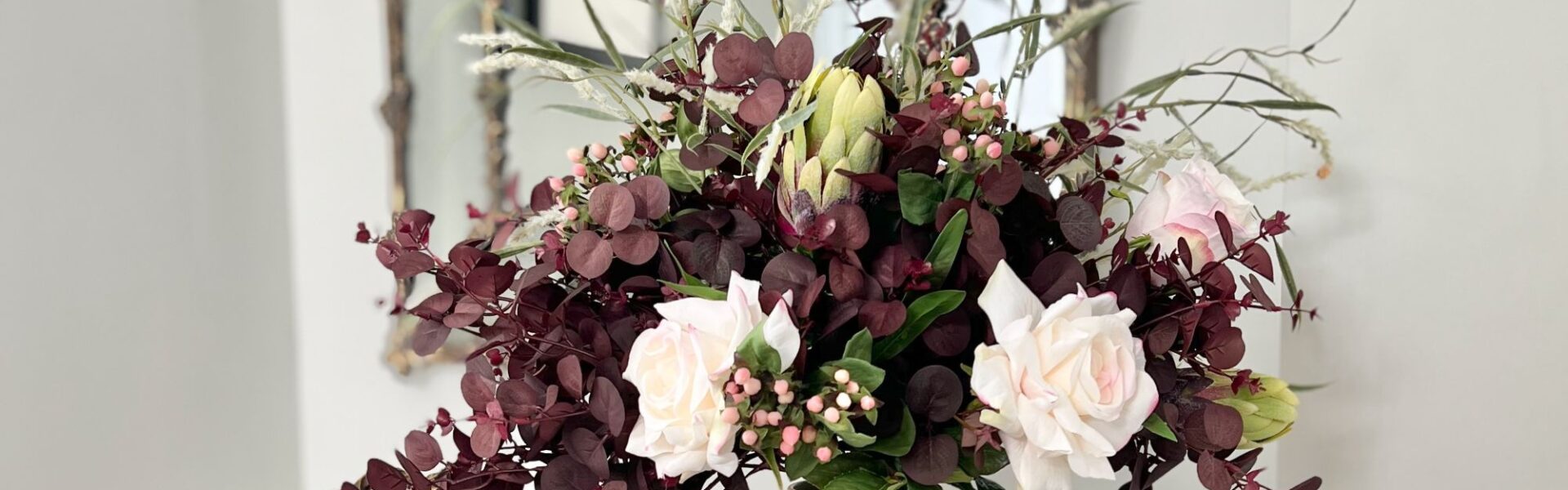A lush autumn floral arrangement in a matte white ceramic vase, featuring deep plum-coloured eucalyptus foliage, pale pink roses, small pink berries, a green protea bud, and wispy ornamental grasses, set against a bright interior with a gold-framed mirror in the background.
