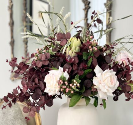 A lush autumn floral arrangement in a matte white ceramic vase, featuring deep plum-coloured eucalyptus foliage, pale pink roses, small pink berries, a green protea bud, and wispy ornamental grasses, set against a bright interior with a gold-framed mirror in the background.