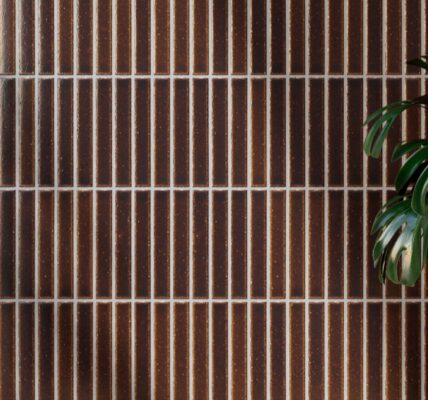 Vertically stacked Kaji Border Japanese porcelain tiles in burnt russet brown with light grout, featured behind a potted Monstera plant.