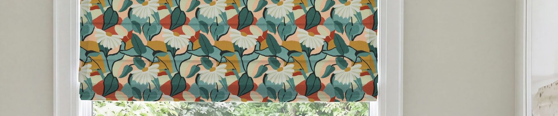A modern Roman blind with a bold botanical pattern of white flowers, dark green and teal leaves, and warm rust-orange and mustard accents, mounted inside a white window frame overlooking green foliage.
