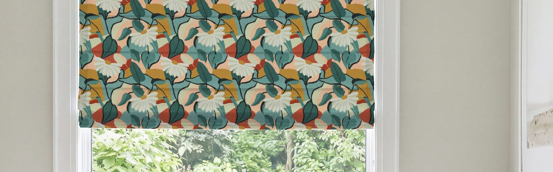 A modern Roman blind with a bold botanical pattern of white flowers, dark green and teal leaves, and warm rust-orange and mustard accents, mounted inside a white window frame overlooking green foliage.