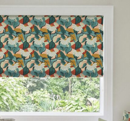 A modern Roman blind with a bold botanical pattern of white flowers, dark green and teal leaves, and warm rust-orange and mustard accents, mounted inside a white window frame overlooking green foliage.
