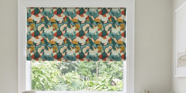 A modern Roman blind with a bold botanical pattern of white flowers, dark green and teal leaves, and warm rust-orange and mustard accents, mounted inside a white window frame overlooking green foliage.