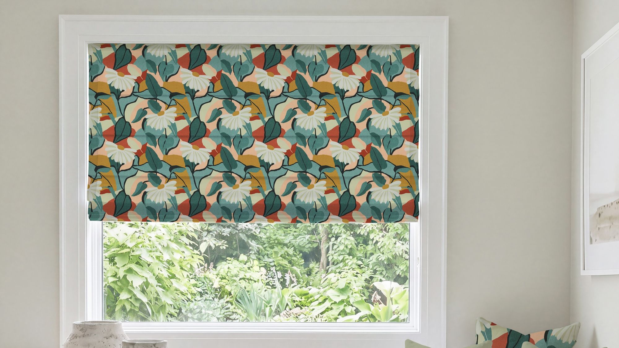 A modern Roman blind with a bold botanical pattern of white flowers, dark green and teal leaves, and warm rust-orange and mustard accents, mounted inside a white window frame overlooking green foliage.