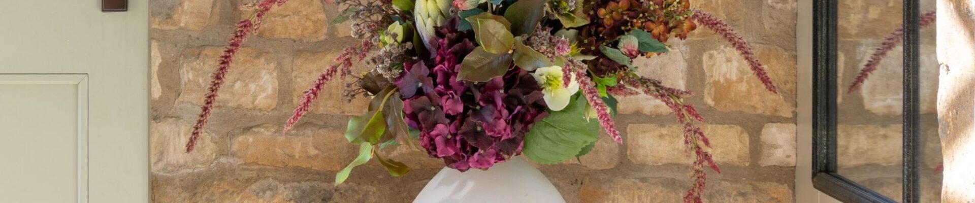 A lush arrangement of dark purple hydrangeas, white flowers, and trailing pink amaranthus in a round white ceramic vase, sitting on a wooden stool against a rustic stone wall.