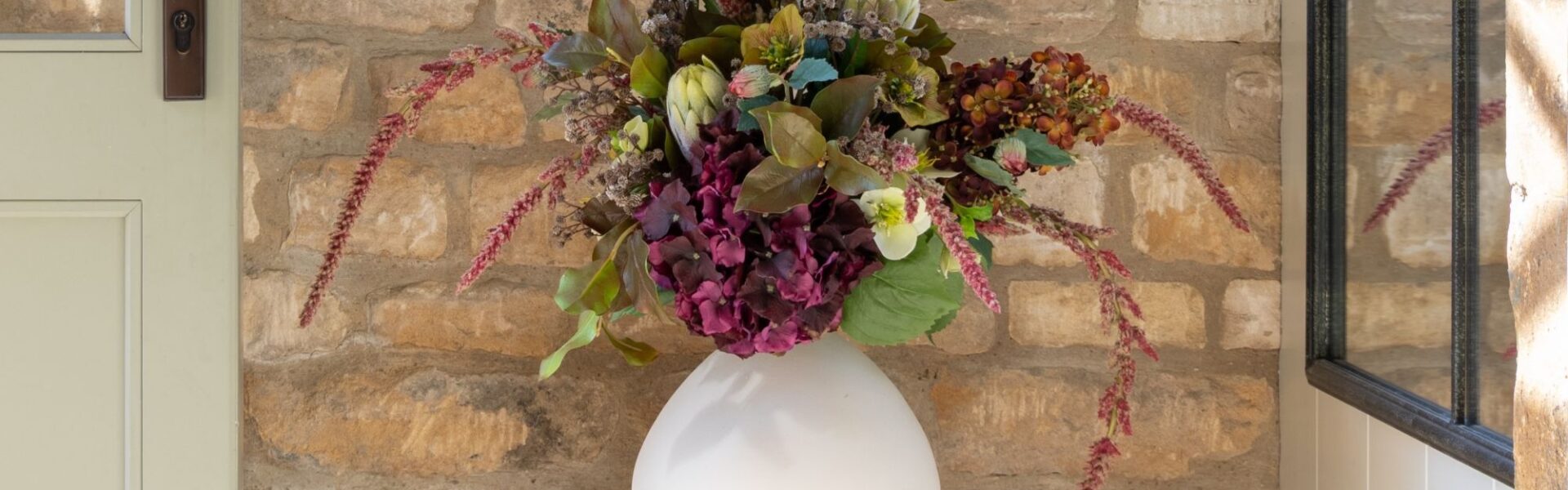 A lush arrangement of dark purple hydrangeas, white flowers, and trailing pink amaranthus in a round white ceramic vase, sitting on a wooden stool against a rustic stone wall.